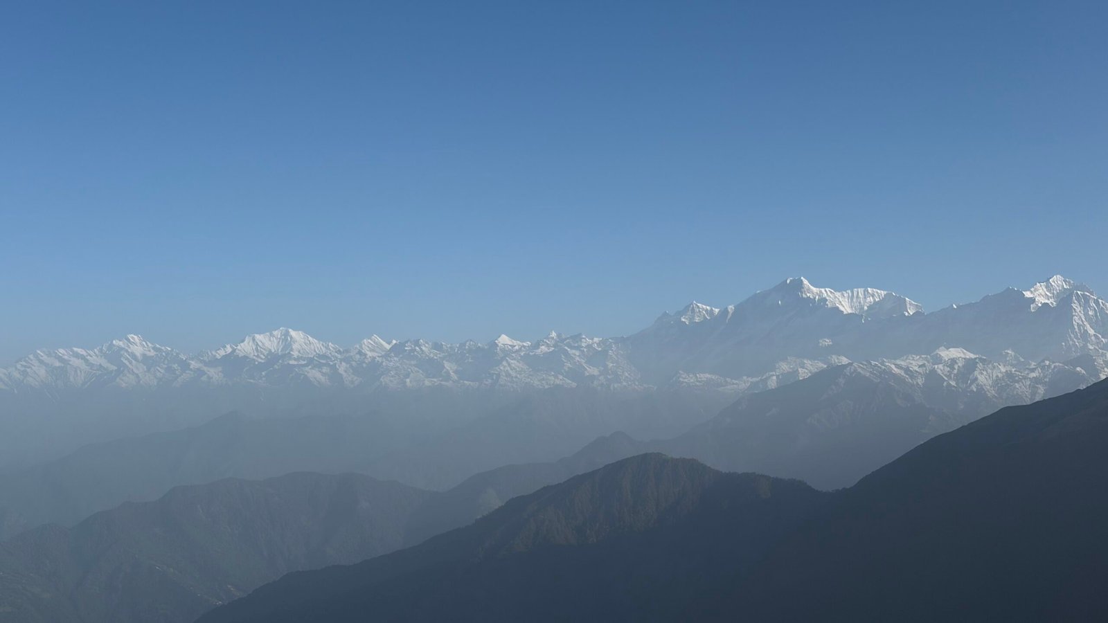 A long line of snow-covered Himalayan peaks rising above hazy layered ridges under a clear blue sky