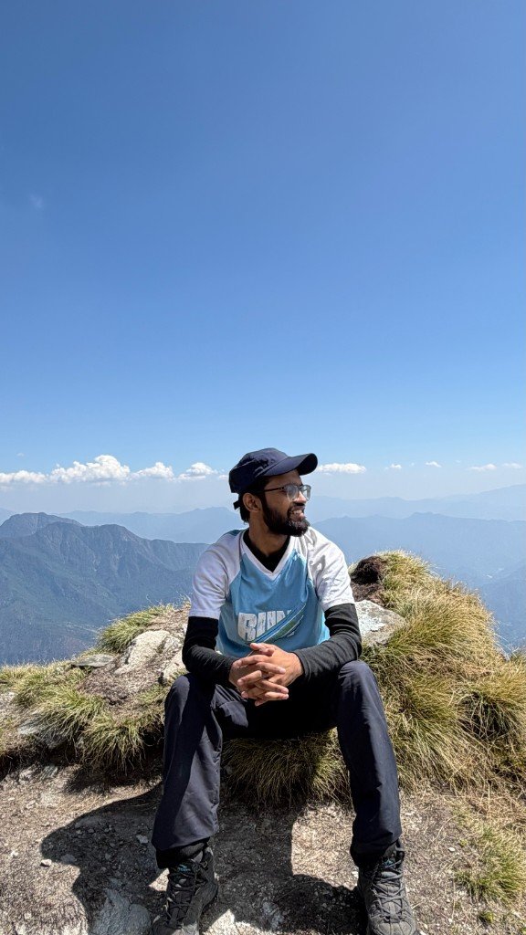 Paras sitting on a rocky Himalayan summit looking out over green ridges under a clear blue sky